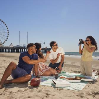 Young couples sit and enjoy pizza on the Atlantic City beach with Steel Pier in background. Woman kneeling holds a camera to snap a picture.