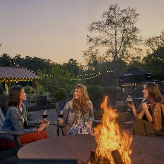 Three women sip wine and chat in an outdoor setting at dusk with a fire pit at Renault Winery outside Atlantic City, NJ.
