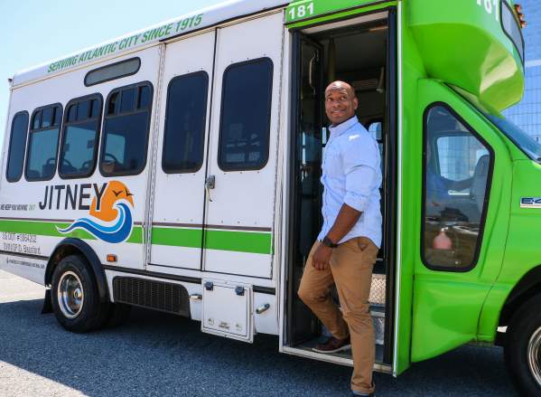 A person stands next to a green and white jitney bus, showcasing its service for Atlantic City, with a clear sky in the background.