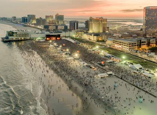 A vast crowd gathers on a beach with concert stage situated on the beach in front of the Atlantic City skyline at sunset.