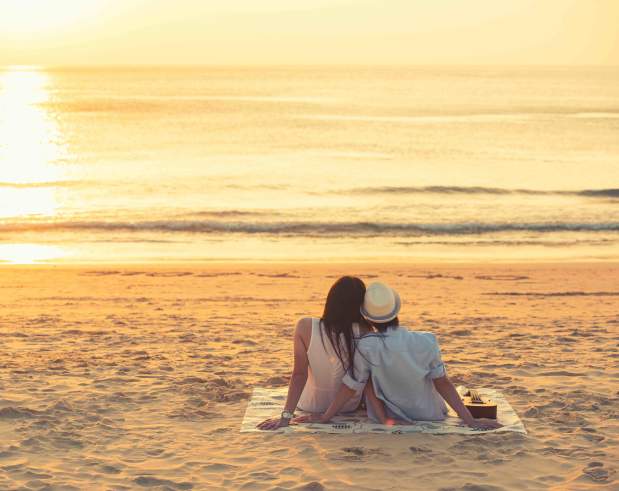A couple sits close to one another on a blanket sitting on the beach watching the sunrise over the calm ocean waves.