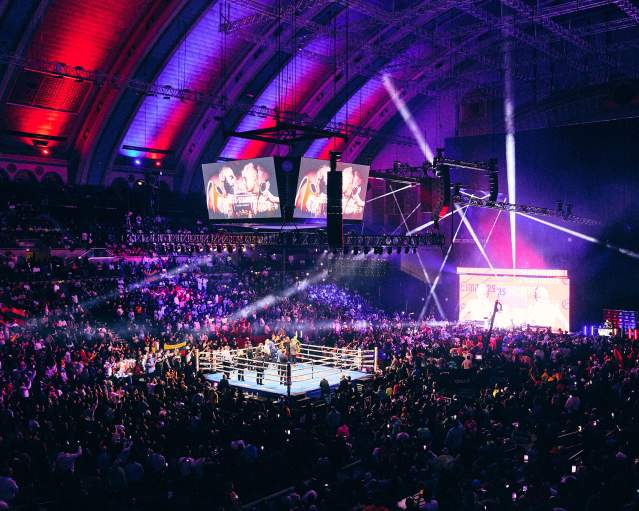 A boxing ring is illuminated in the foreground with a large crowd watching, while the background is filled with a dazzling light show and screens displaying images.