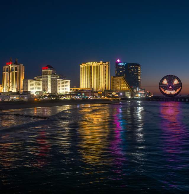 A dark ocean reflects the illuminated skyline of Atlantic City, featuring a giant jack-o'-lantern on a pier in the background.