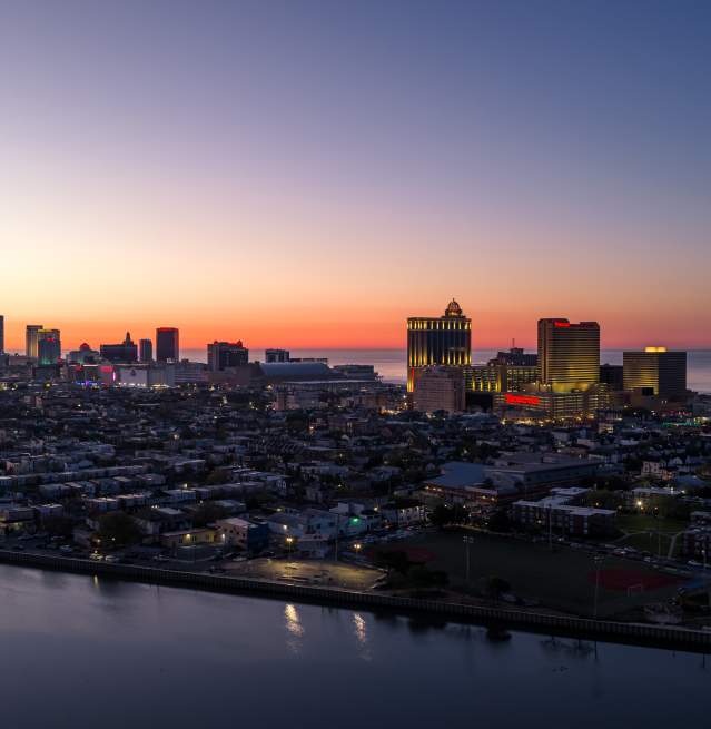 A calm body of water in the foreground leads to the Atlantic City skyline illuminated by the soft glow of sunrise in the background.