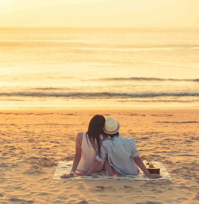 A couple sits close to one another on a blanket sitting on the beach watching the sunrise over the calm ocean waves.
