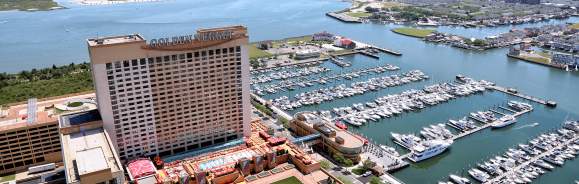 A large casino hotel with a marina full of boats sits on the waterfront, with a distant coastline and ocean under a clear blue sky.