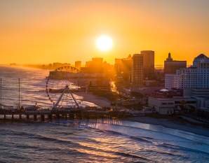 The setting sun shines over the Atlantic City skyline with the Steel Pier and ocean waves in the foreground.