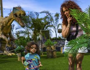 A young child with a toy dinosaur stands on a grassy field in front of a large dinosaur animatronic, while a woman peeks from behind a fern with her finger to her lips.