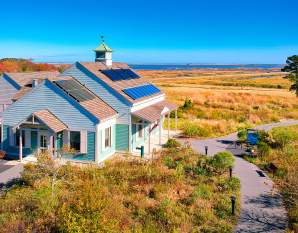 A light blue building with solar panels sits in the foreground, surrounded by dry grass and a winding path, with a vast, calm body of water and a clear blue sky in the background.