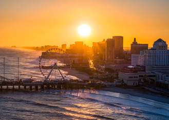 The setting sun shines over the Atlantic City skyline with the Steel Pier and ocean waves in the foreground.