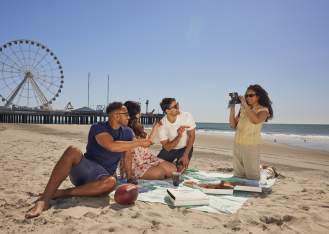 Young couples sit and enjoy pizza on the Atlantic City beach with Steel Pier in background. Woman kneeling holds a camera to snap a picture.
