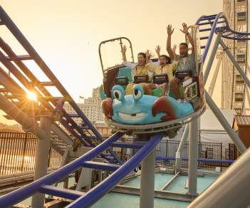 A family rides all new Crazy Crab Coaster with arms raised high descending a coaster section as sun sets behind the Steel Pier on the Atlantic City Boardwalk.
