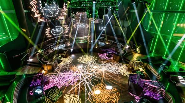 A vibrant nightclub with illuminated dance floors and stage lighting in the foreground, set against a backdrop of green neon lights and a circular truss system in the background.
