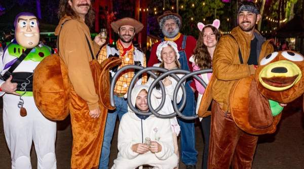 A group of friends in Toy Story costumes pose in front of a festive, tree-lined background with colorful lights.