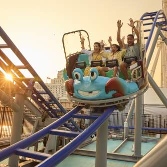 A family rides all new Crazy Crab Coaster with arms raised high descending a coaster section as sun sets behind the Steel Pier on the Atlantic City Boardwalk.