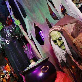Two spooky witch decorations, one with green skin and a cauldron in the foreground, are set against a dimly lit background with people and colorful lights.