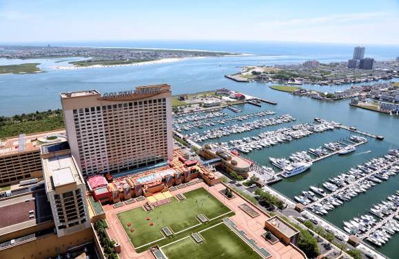 A large casino hotel with a marina full of boats sits on the waterfront, with a distant coastline and ocean under a clear blue sky.