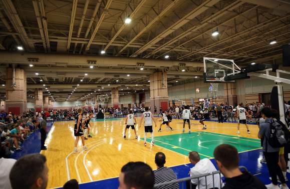 Players compete on a brightly lit basketball court in the foreground, while a large crowd watches from the bleachers in the background.