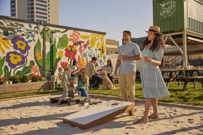 A couple enjoys a sunny day at Cardinal outdoor space with a colorful mural and sandpit in the foreground, while a cityscape and a shipping container garden are visible in the background.