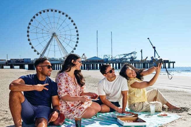 People seated taking a selfie enjoying Boardwalk food on an Atlantic City beach with Steel Pier's The Wheel and shoreline in view.