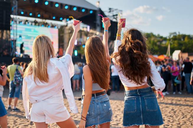 3 women holding up drinks in front of a stage set up having a great time at a beach festival.