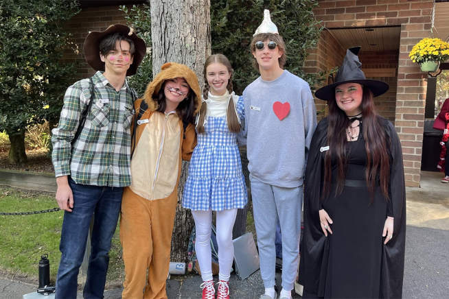 Five friends in costumes, including a scarecrow, bear, Dorothy, Tin Man, and witch, pose in front of a building with a tree and foliage.