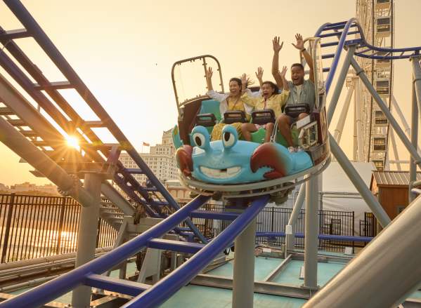 A family rides all new Crazy Crab Coaster with arms raised high descending a coaster section as sun sets behind the Steel Pier on the Atlantic City Boardwalk.