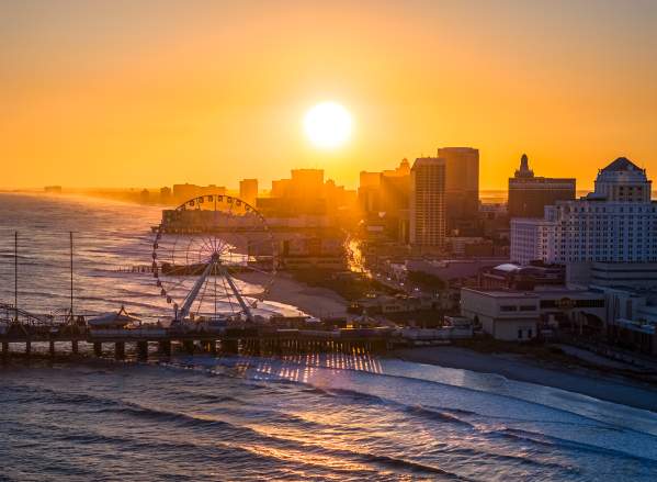 The setting sun shines over the Atlantic City skyline with the Steel Pier and ocean waves in the foreground.
