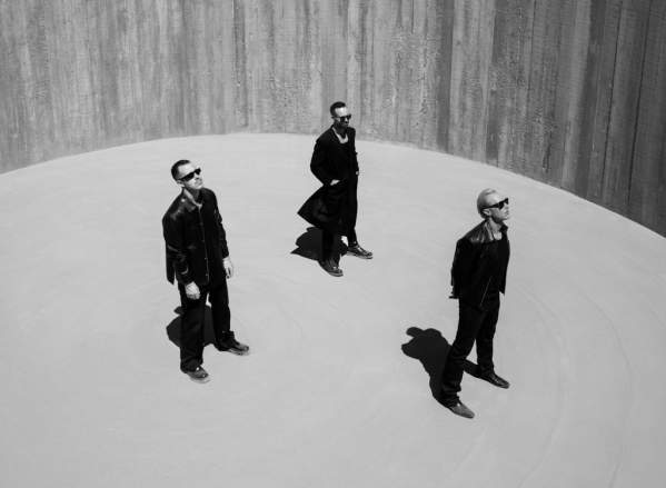 Three men stand in a circular, sandy pit with textured, vertical walls rising behind them.