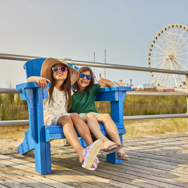 Two kids with sunglasses enjoy a break in a decoratively painted Adirondack chair on the Atlantic City Boardwalk with Steel Pier's The Wheel in background.