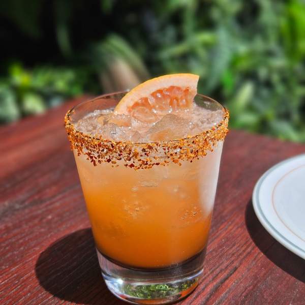 A grapefruit-garnished cocktail with a spiced rim sits on a wooden table in front of a blurred green foliage background.