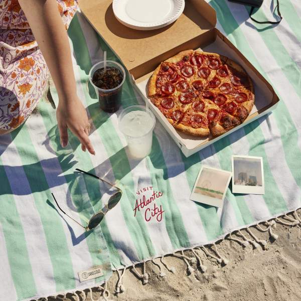Young woman sits on beach blanket with snapshot photos, sunglasses, pepperoni pizza on sunny day on the Atlantic City beach.