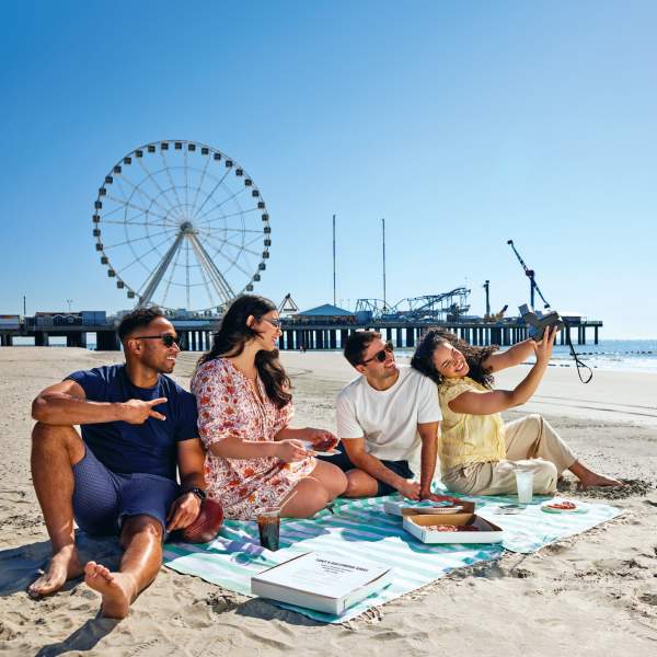 People seated taking a selfie enjoying Boardwalk food on an Atlantic City beach with Steel Pier's The Wheel and shoreline in view.