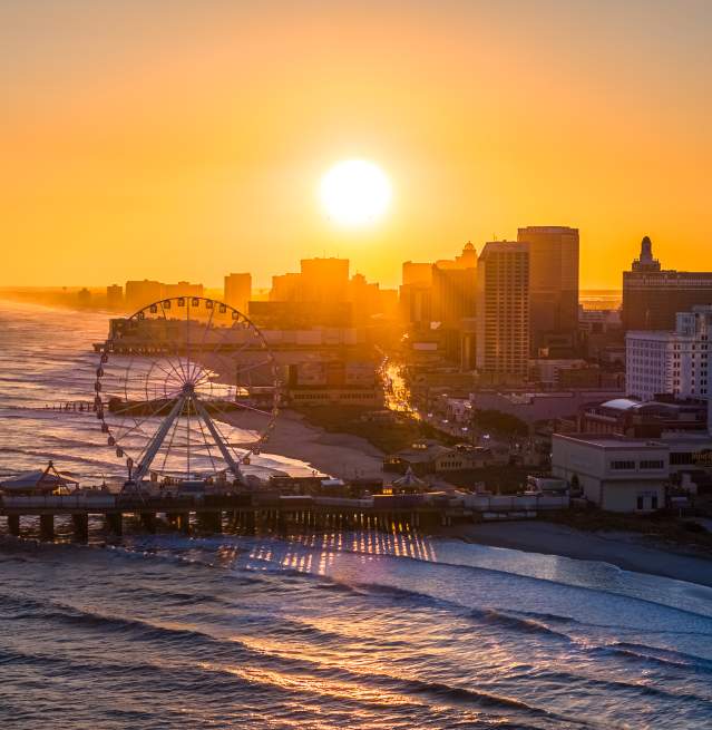 The setting sun shines over the Atlantic City skyline with the Steel Pier and ocean waves in the foreground.