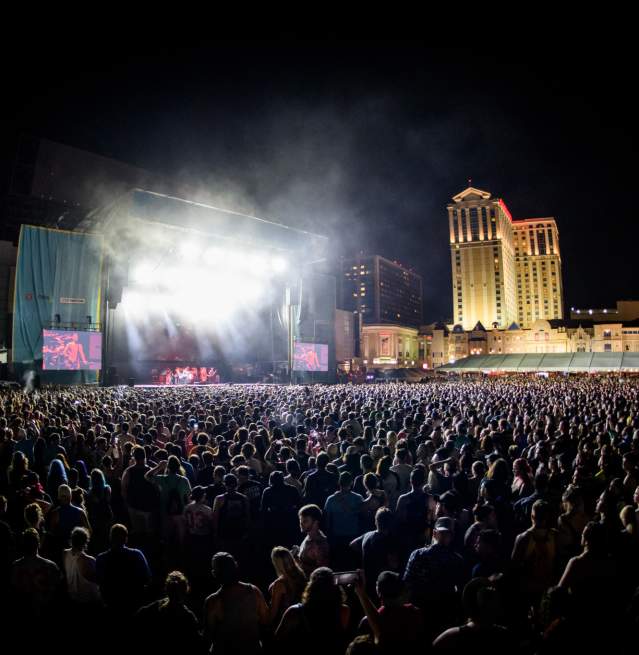 A massive crowd gathers in the foreground for an outdoor beach concert in Atlantic City, with a brightly lit stage and a tall, illuminated building in the background.