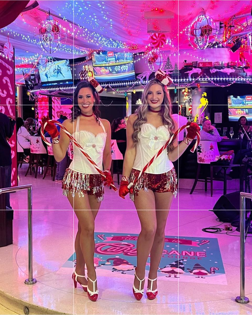 Two women dressed in themed attire holding large candy canes in front of the Candy Cane Lounge Pop Up Experience at Bar One inside Resorts Casino Hotel.