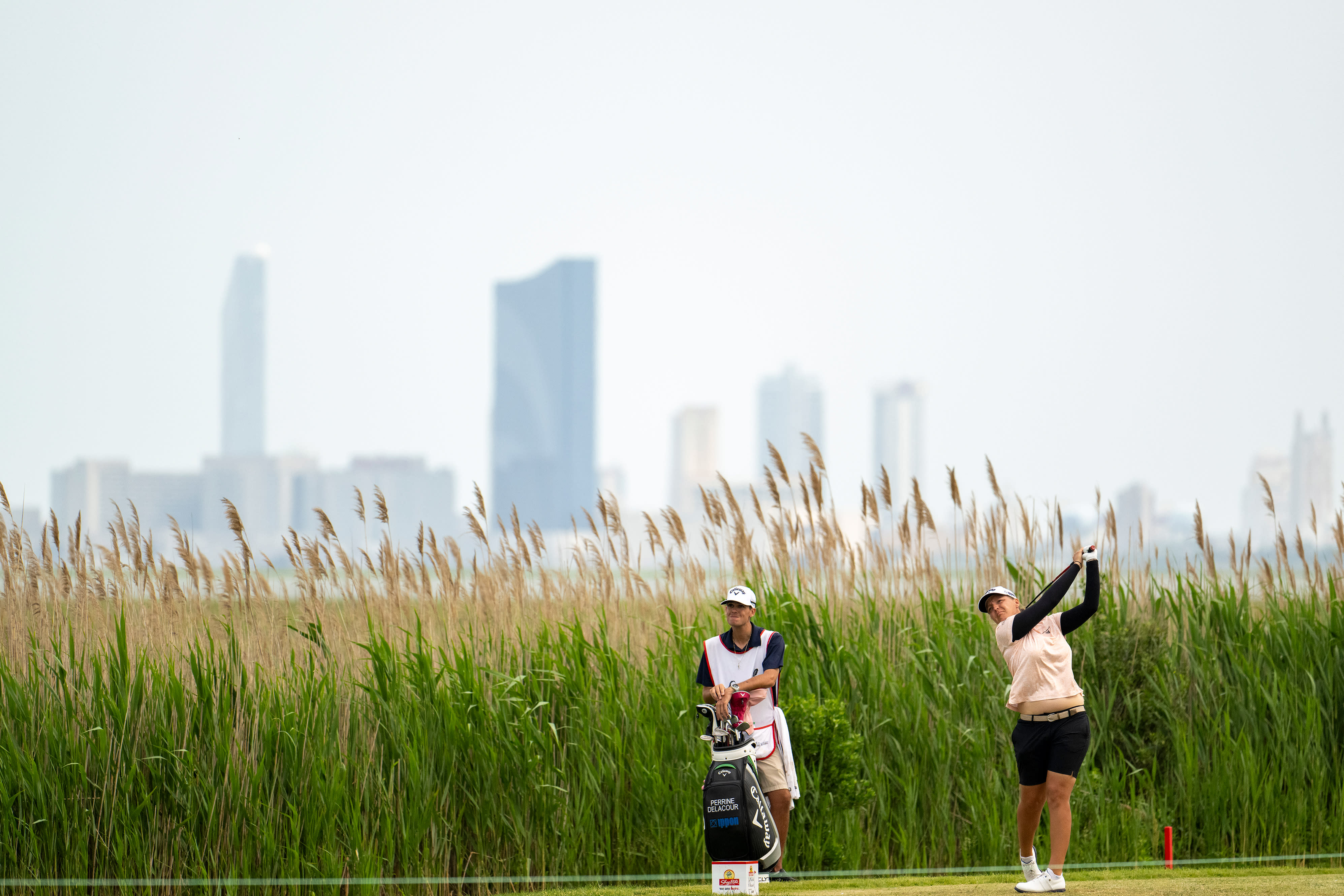 LPGA professional tees off on the Bay Course at Seaview with marsh reeds and Atlantic City in distance.
