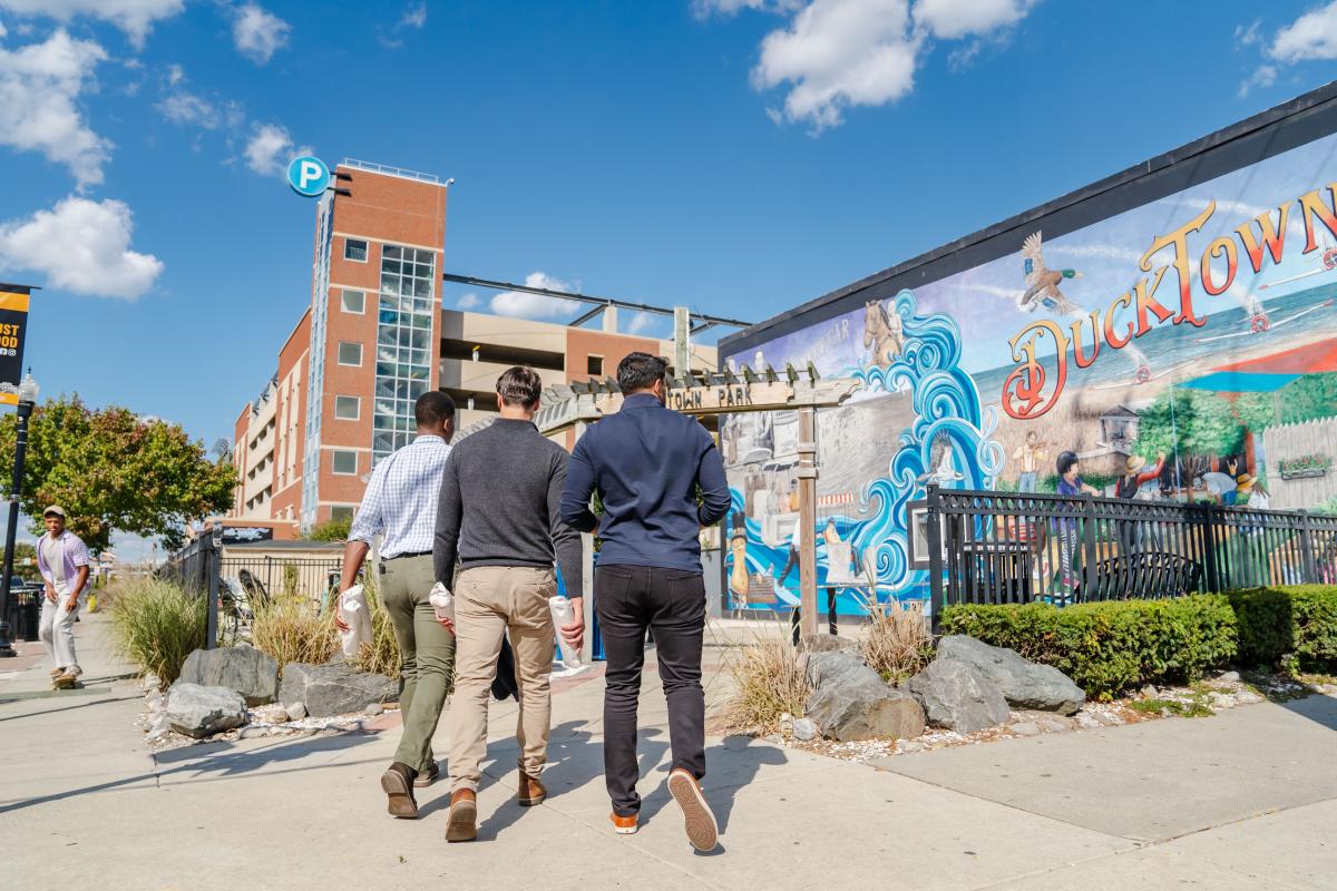 Three guys walk in the Ducktown Neighborhood in Atlantic City as a person on a skateboard rides by with Ducktown Mural and Wave Parking Garage in view.