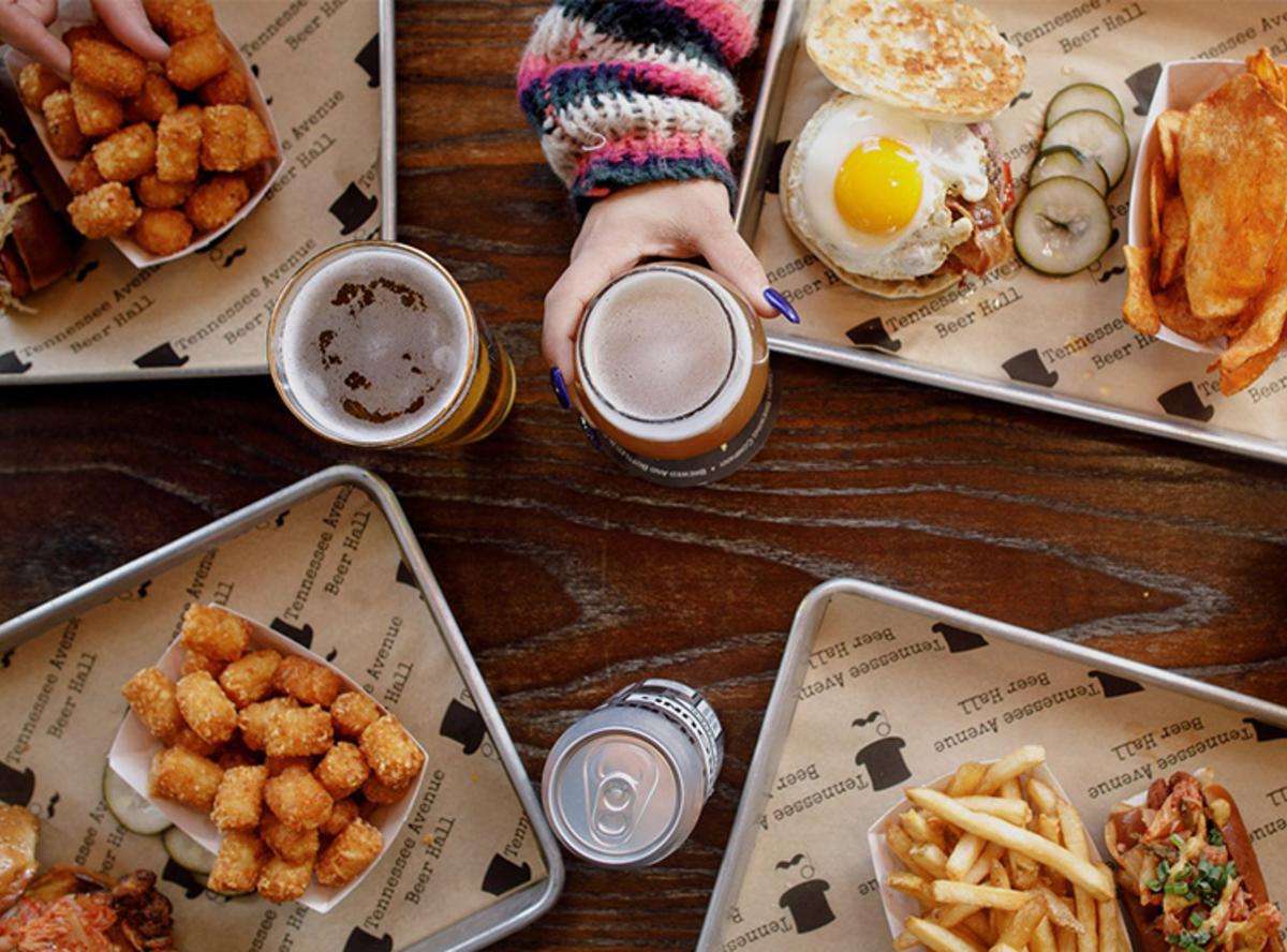 Aluminum trays filled with Tennessee Avenue Beer Hall fare rests on a wooden table with glasses of craft beer and one canned beer.