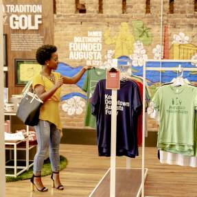 A person examines t-shirts featuring Augusta-themed designs in a colorful store with a mural depicting local history and attractions.
