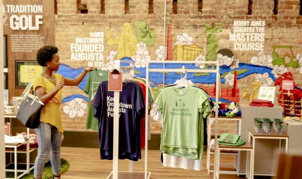A person examines t-shirts featuring Augusta-themed designs in a colorful store with a mural depicting local history and attractions.