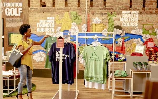 A person examines t-shirts featuring Augusta-themed designs in a colorful store with a mural depicting local history and attractions.