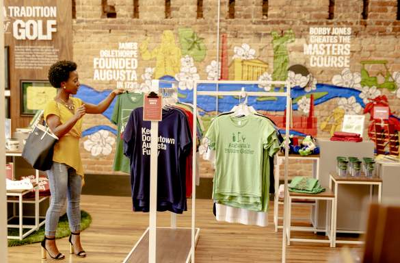 A person examines t-shirts featuring Augusta-themed designs in a colorful store with a mural depicting local history and attractions.