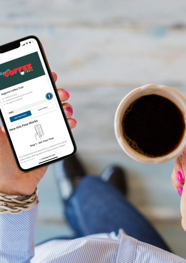 Woman with bright pink nails holds a coffee and mobile phone showing the Augusta Coffee Trail pass.