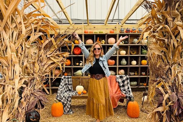 A woman in a denim jacket and long skirt stands joyfully among decorative pumpkins and corn stalks, celebrating autumn.