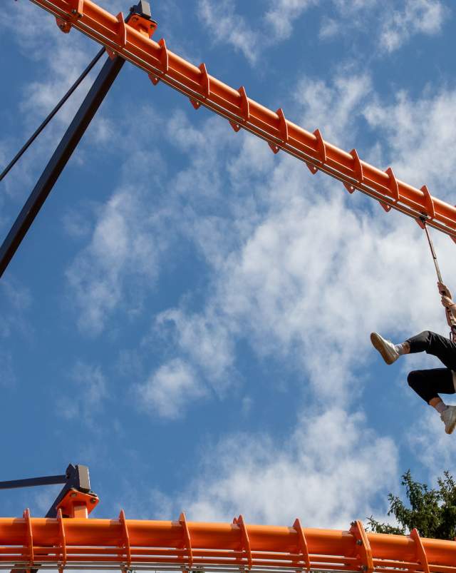 A person swings from a zipline high above the ground at Binder Park Zoo, set against a vivid blue sky with scattered clouds.