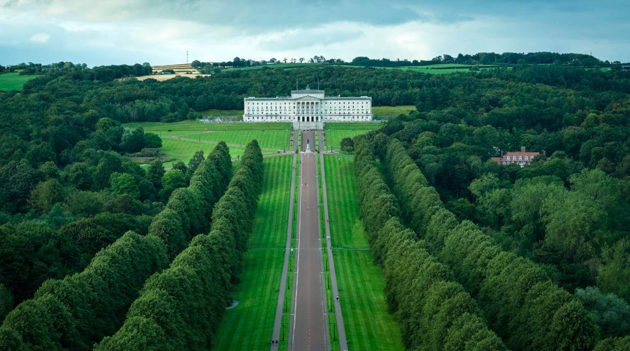 A grand white building stands at the end of a long, manicured lawn lined with trees, surrounded by rolling green hills and a cloudy sky.