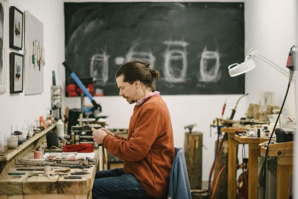 A man in an orange sweater works at a jewelry bench in the foreground, with a large chalkboard and studio equipment in the background.