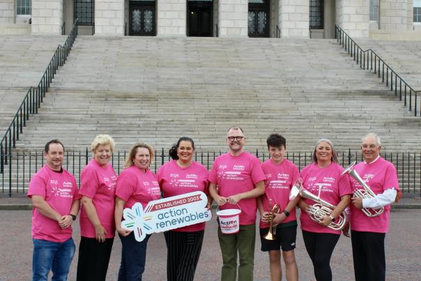 A group of people in pink shirts stand on a paved area in front of a grand building with large stone steps.