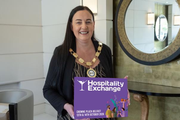 A woman in a chain of office holds a purple sign for "Hospitality Exchange" in front of a large ornate mirror.